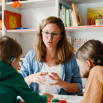 Kindergarten; zwei Kinder sitzen mit einer erwachsenen Person an einem Tisch und spielen ein Spiel. Dabei werden Gebärden eingesetzt. 