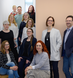 Eine Gruppe von zwölf Erwachsenen steht und sitzt für ein gemeinsames Foto im Innenbereich. Die Personen sind freundlich und entspannt, sie schauen in die Kamera und lächeln. Die Gruppe besteht aus zehn Frauen und zwei Männern. Im Vordergrund sitzen zwei Frauen, die restlichen Personen stehen in zwei Reihen – einige auf einer Treppe, andere daneben vor einer hellen Wand. Die Kleidung ist überwiegend in gedeckten Farben wie Schwarz, Grau, Blau und Weiß gehalten.